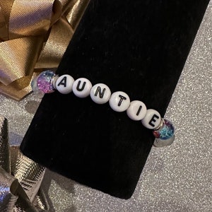 Puede incluir: Una pulsera de cuentas blancas que deletrea "AUNTIE" en letras negras. La pulsera tiene dos cuentas iridiscentes y brillantes en cada extremo. La pulsera se muestra sobre una superficie negra con un fondo dorado y plateado.