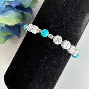 May include: A beaded bracelet spelling "NICE" with white letter beads and turquoise accent beads. The bracelet is displayed on a black velvet surface, with a blue and white flower in the background.