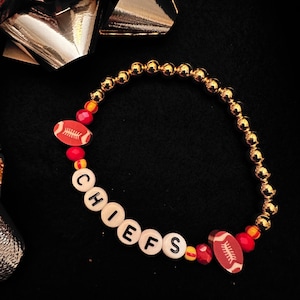 Könnte beinhalten: Ein goldfarbenes Perlenarmband mit dem Wort "CHIEFS" aus weißen Buchstabenperlen. Das Armband enthält zwei fußballförmige Perlen und rote und gelbe Akzentperlen. Das Armband liegt auf einem schwarzen Hintergrund mit einer silbernen Schleife.