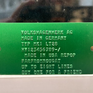 May include: A green metal plate with rounded corners and two holes. The plate has embossed text that reads "VOLKSWAGENVERK AG MADE IN GERMANY TYP MK1 LT28 MV123456789-/ MADE IN USA REPOP FARFROMNOUGAT UP TO EIGHT LINES BUY ONE FOR A FRIEND."