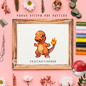 Op de afbeelding: Kruissteekpatroon van een rood en oranje Pokémon-personage, Charmander, met een vlammende staart. Het patroon bevindt zich in een houten frame op een roze achtergrond. De tekst "CROSS STITCH PDF PATTERN" staat bovenaan het frame en "CRAFTPATTERNHUB" staat onderaan het frame.