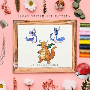 May include: Cross stitch pattern featuring three Pokemon characters: Charizard, a red and orange dragon, and two blue and white Dratini. The pattern is framed in a wooden frame and the text "CROSS STITCH PDF PATTERN" and "CRAFTPATTERNHUB" is visible.