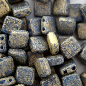May include: A close-up of a pile of small, square, gold and blue glass beads. The beads have a textured, speckled surface.