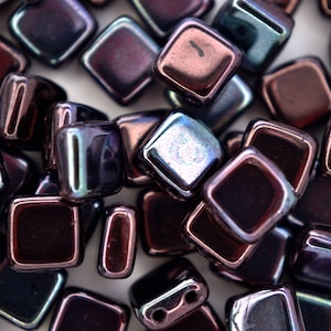 May include: A collection of iridescent square beads in shades of purple, brown, and blue. The beads have a glossy finish and are arranged closely together, showcasing their geometric shape and reflective surfaces. Ideal for jewelry making.