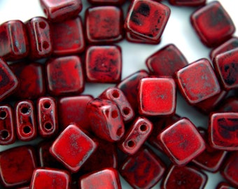 May include: A close-up of red square beads with black speckles. The beads are made of glass and have a matte finish.