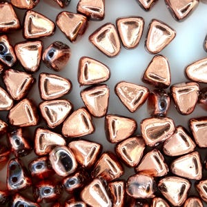 May include: A collection of copper-coloured, triangular beads with a metallic sheen. The beads have a flat back and a rounded, slightly raised front. Some beads have a hole for stringing. The beads are scattered on a white surface.