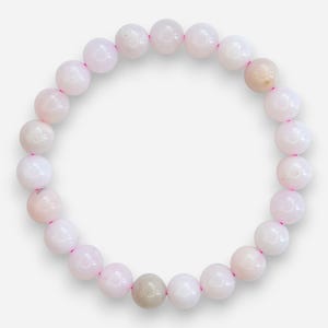 May include: A beaded bracelet featuring round, light pink beads strung on a pink elastic cord. The bracelet has a few beads that are a light tan color. The beads are smooth and evenly sized, creating a simple, elegant accessory.