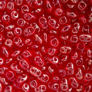 May include: Close-up of numerous small, translucent red beads. Each bead has a rounded form with a central hole, producing a textured appearance. The image accentuates the vivid colour and intricate details of the beads, which could be used for jewellery.