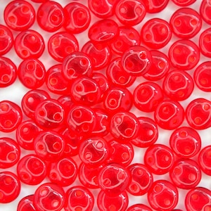 May include: A close-up of a pile of red glass beads with two holes each. The beads are round and have a smooth, shiny finish.