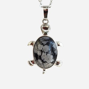 Può includere: Una collana con pendente argentato a forma di tartaruga. Il pendente presenta una pietra ovale nera e grigia incastonata in una cornice di metallo argentato. La testa e le pinne della tartaruga sono anch'esse argentate. La collana è appesa a una delicata catena d'argento.