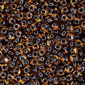 Può includere: Primo piano di un mucchio di piccole perline quadrate. Le perline sono un mix di colori marrone scuro e ambra, con una finitura lucida. Le perline sono sparse, creando una superficie strutturata, ideale per la creazione di gioielli.