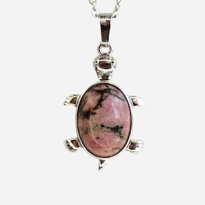 May include: A silver pendant necklace featuring a turtle design. The pendant has a pink and black oval-shaped stone set in a silver turtle frame. The necklace is on a white background.