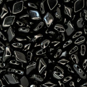 Puede incluir: Primer plano de un montón de cuentas negras brillantes. Las cuentas son una mezcla de formas de diamante y ovaladas, cada una con dos agujeros para ensartar. La superficie reflectante de las cuentas crea reflejos.