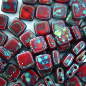 May include: A close-up of red and blue square-shaped glass beads with a mottled, marbled effect. The beads are scattered on a white surface.