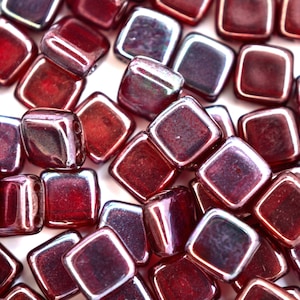 May include: A close-up of a group of small, square, iridescent glass beads. The beads are a deep red color with a slight purple hue. They are arranged in a random pattern on a white background.