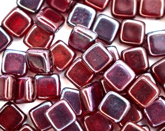 May include: A close-up of a group of small, square, iridescent glass beads. The beads are a deep red color with a slight purple hue. They are arranged in a random pattern on a white background.