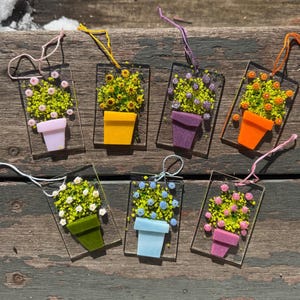 May include: A collection of seven rectangular glass ornaments, each featuring a colorful flower arrangement in a pot. The pots are various colors, including pink, yellow, orange, and green. Each ornament has a string for hanging.