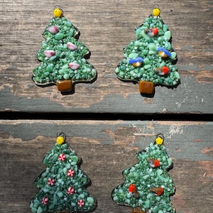 May include: Four handmade Christmas tree ornaments. Each tree is green with a brown trunk and a yellow loop for hanging. The trees are decorated with small birds, candies, and other colorful accents. The ornaments are on a weathered wooden surface.