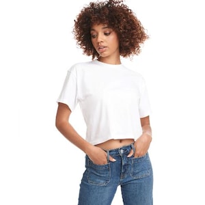 May include: A white, cropped t-shirt with short sleeves. The model is wearing blue denim jeans with pockets. The background is plain white.