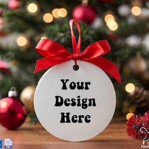 May include: White round Christmas ornament with a red ribbon and the text "Your Design Here" on a wooden surface with a Christmas tree in the background.