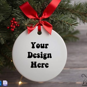 May include: White round Christmas ornament with a red ribbon hanging from a green pine tree branch. The ornament has the text "Your Design Here" printed on it.