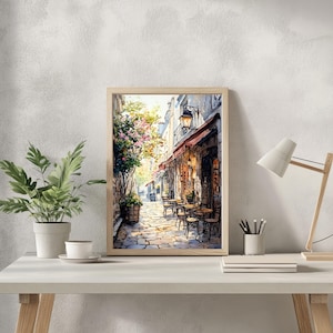 May include: A watercolor painting of a Parisian street scene with a cafe on the corner. The cafe has a red awning and tables and chairs set up outside. The street is cobblestone and there are buildings on both sides. The painting is framed in a light wood frame.