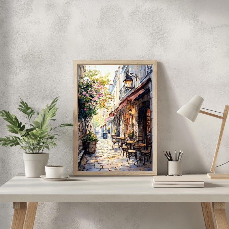 May include: A watercolor painting of a Parisian street scene with a cafe on the corner. The cafe has a red awning and tables and chairs set up outside. The street is cobblestone and there are buildings on both sides. The painting is framed in a light wood frame.