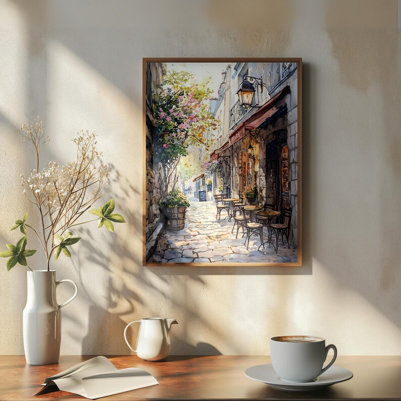 May include: A watercolor painting of a narrow street in a European city. The street is lined with buildings and there are tables and chairs set up outside a cafe. The painting is done in a realistic style and the colors are muted and soft.