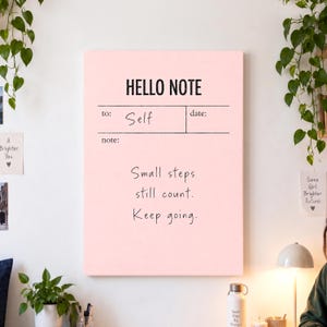 May include: A rectangular light pink wall art piece featuring the black text HELLO NOTE at the top. Below, lines for to: Self and date: are printed. The centre displays the black handwritten text: Small steps still count. Keep going.