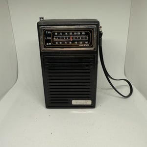 Puede incluir: Una radio portátil negra vintage con una cara plateada. La radio tiene un dial FM y AM, con los números 55 a 160. Una correa negra está sujeta al lateral. Las palabras "SOLID STATE" están impresas en la cara.