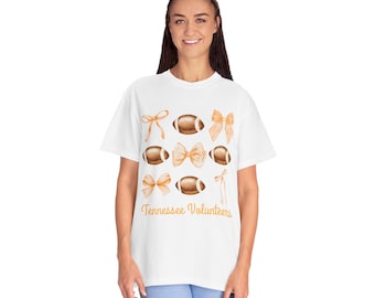 May include: White t-shirt featuring watercolor illustrations of footballs and bows in shades of brown and orange. The text "Tennessee Volunteers" is printed in orange script below the graphics. The shirt is a casual, unisex style.