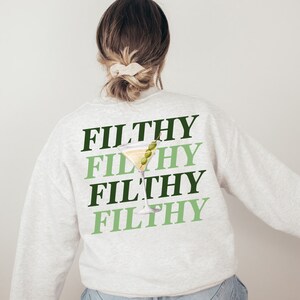 May include: A light gray sweatshirt with the word "FILTHY" repeated four times in black and green lettering. A green olive and a martini glass are superimposed on the word "FILTHY".