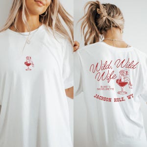 Op de afbeelding: Wit T-shirt met een rode afbeelding van een vrouw in een cocktailglas op de voorkant. De achterkant van het shirt heeft de tekst "Wild, Wild Wife" en "Blake's Bachelorette" met "Jackson Hole, WY" eronder.