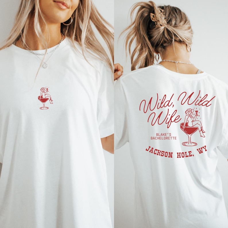 Op de afbeelding: Wit T-shirt met een rode afbeelding van een vrouw in een cocktailglas op de voorkant. De achterkant van het shirt heeft de tekst "Wild, Wild Wife" en "Blake's Bachelorette" met "Jackson Hole, WY" eronder.