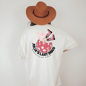 May include: White t-shirt with a pink and black graphic of a disco ball with legs wearing cowboy boots. The text "Julia's Last Disco Nashville 2024" is printed on the shirt.