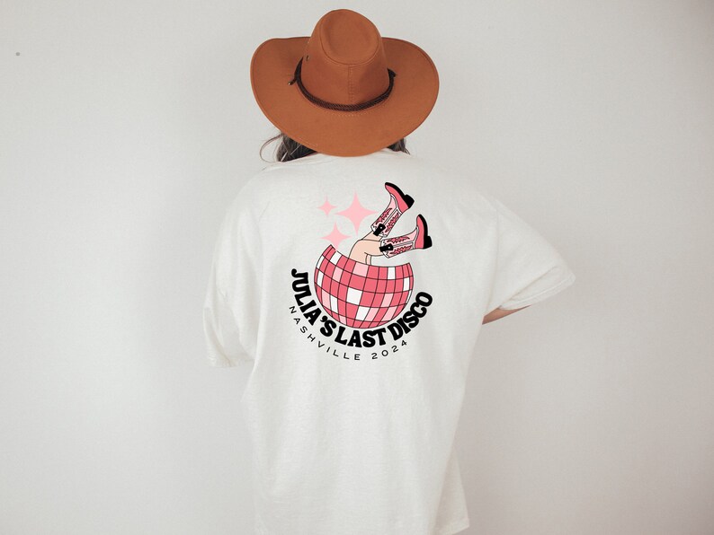 May include: White t-shirt with a pink and black graphic of a disco ball with legs wearing cowboy boots. The text "Julia's Last Disco Nashville 2024" is printed on the shirt.