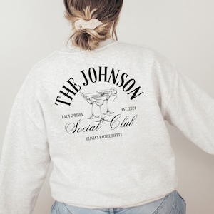 K&ouml;nnte beinhalten: Ein grauer Pullover mit schwarzem Text, der "The Johnson Social Club, Palm Springs, Est. 2024, Olivia's Bachelorette" lautet und eine Grafik von zwei Martini-Gl&auml;sern.