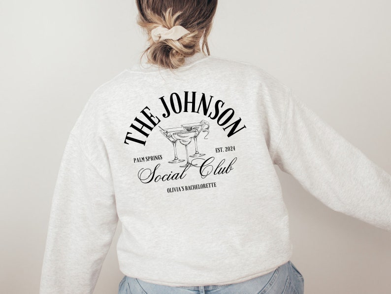 K&ouml;nnte beinhalten: Ein grauer Pullover mit schwarzem Text, der "The Johnson Social Club, Palm Springs, Est. 2024, Olivia's Bachelorette" lautet und eine Grafik von zwei Martini-Gl&auml;sern.