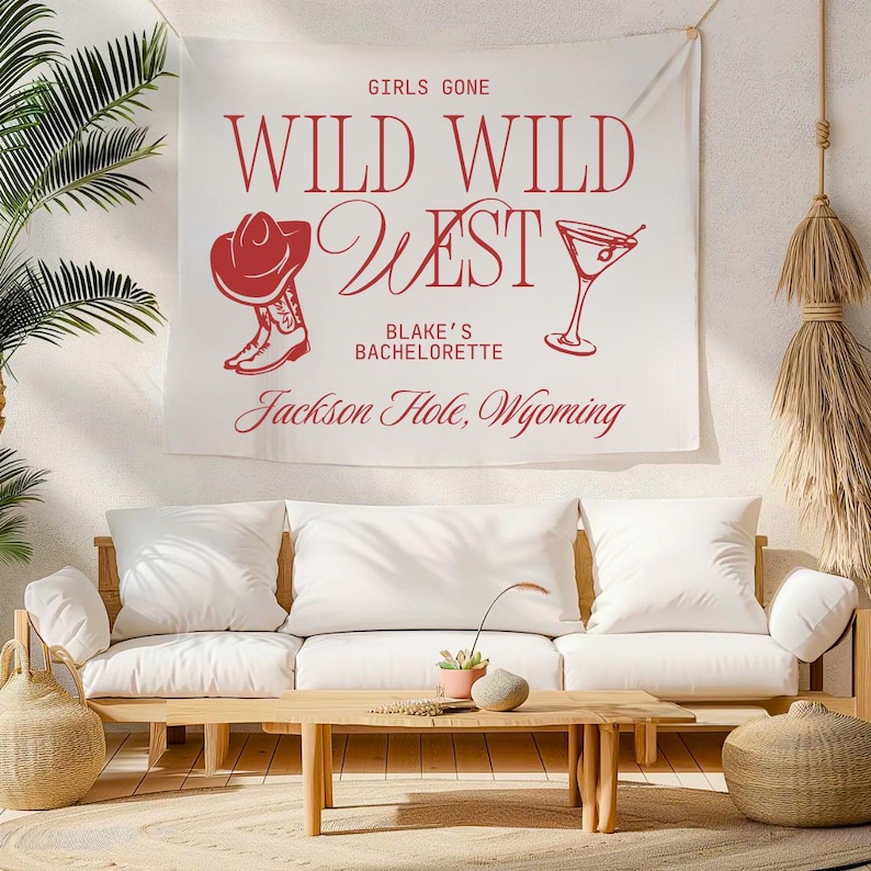 Op de afbeelding: Een witte stoffen wandkleed met rode tekst en afbeeldingen met de tekst "GIRLS GONE WILD WILD WEST" met een cowboyhoed, laars en martiniglas. Daaronder staat "Blake's Bachelorette Jackson Hole, Wyoming".