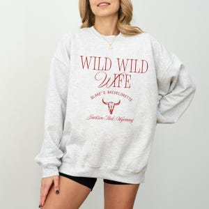 Op de afbeelding: Een lichtgrijs sweatshirt met de tekst "WILD WILD WIFE" in bordeauxrood. Daaronder staat "Blake's Bachelorette" en een grafische afbeelding van een schedel van een stier. De tekst "Jackson Hole, Wyoming" staat onderaan.
