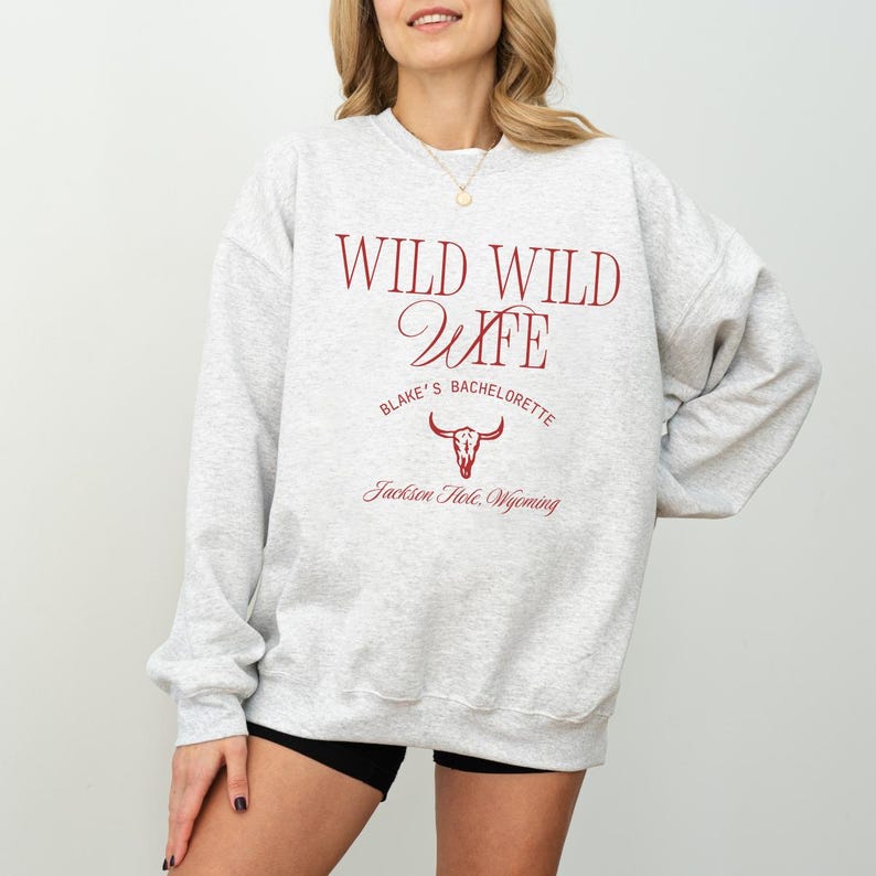 Op de afbeelding: Een lichtgrijs sweatshirt met de tekst "WILD WILD WIFE" in bordeauxrood. Daaronder staat "Blake's Bachelorette" en een grafische afbeelding van een schedel van een stier. De tekst "Jackson Hole, Wyoming" staat onderaan.