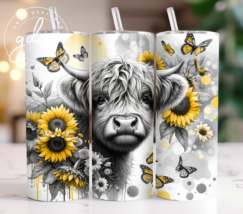 May include: A white tumbler with a black and white illustration of a Highland cow with yellow sunflowers and butterflies. The tumbler has a clear lid and a straw.