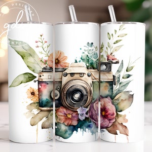 May include: A white tumbler with a watercolor illustration of a vintage camera surrounded by flowers and leaves. The camera is brown and gold with a black lens. The flowers are pink, purple, and orange.