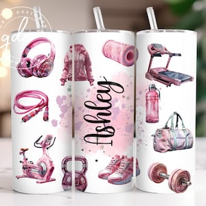 Puede incluir: Un vaso blanco con un tema de fitness rosa. El vaso presenta ilustraciones de equipos de entrenamiento como auriculares, una cinta de correr y pesas. El nombre "Ashley" está escrito en escritura negra sobre una salpicadura de acuarela rosa.