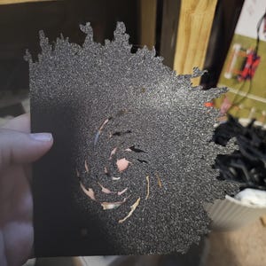 May include: A black stencil with a spiral design cut out. The stencil is made of a textured material and has a rough, unfinished look.