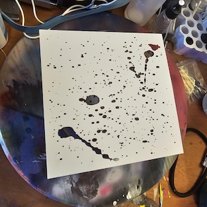 May include: A white square paper with black ink splatters and drips. The ink varies in size, creating a dynamic abstract design. The artwork is likely for decorative purposes or artistic expression.