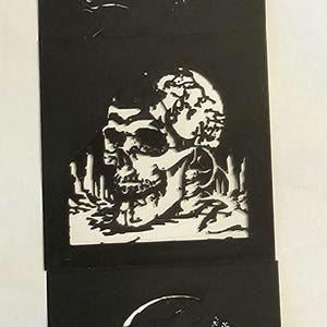 May include: A black paper art piece featuring three cut-out images. The top image is a silhouette of a person, the middle is a skull, and the bottom is a skull. The cut-outs are white.