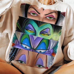 May include: Beige sweatshirt featuring a graphic of three villainous female characters' eyes. The top character has green eyes and dark makeup, the middle has yellow eyes and green skin, and the bottom has blue eyes and purple eyeshadow.