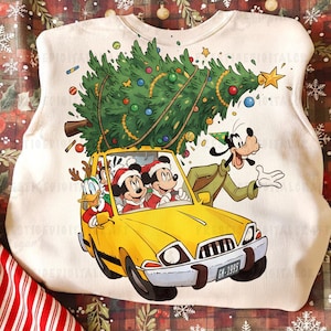 Peut inclure: Un sweat-shirt blanc avec un motif festif. Il représente une voiture jaune transportant un sapin de Noël, conduite par Mickey et Minnie Mouse, avec Donald Duck et Dingo. Le sapin est orné de décorations. La plaque d'immatriculation de la voiture indique "GM-1935".