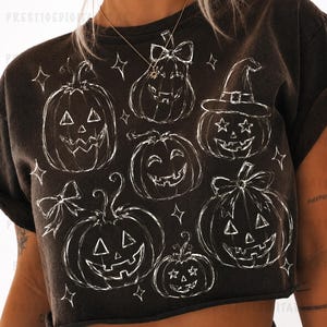 May include: A dark brown, short-sleeved, cropped t-shirt featuring a white, hand-drawn style graphic print of six jack-o'-lanterns. The pumpkins are decorated with various elements including bows, a witch hat, and star-shaped eyes, surrounded by small stars.